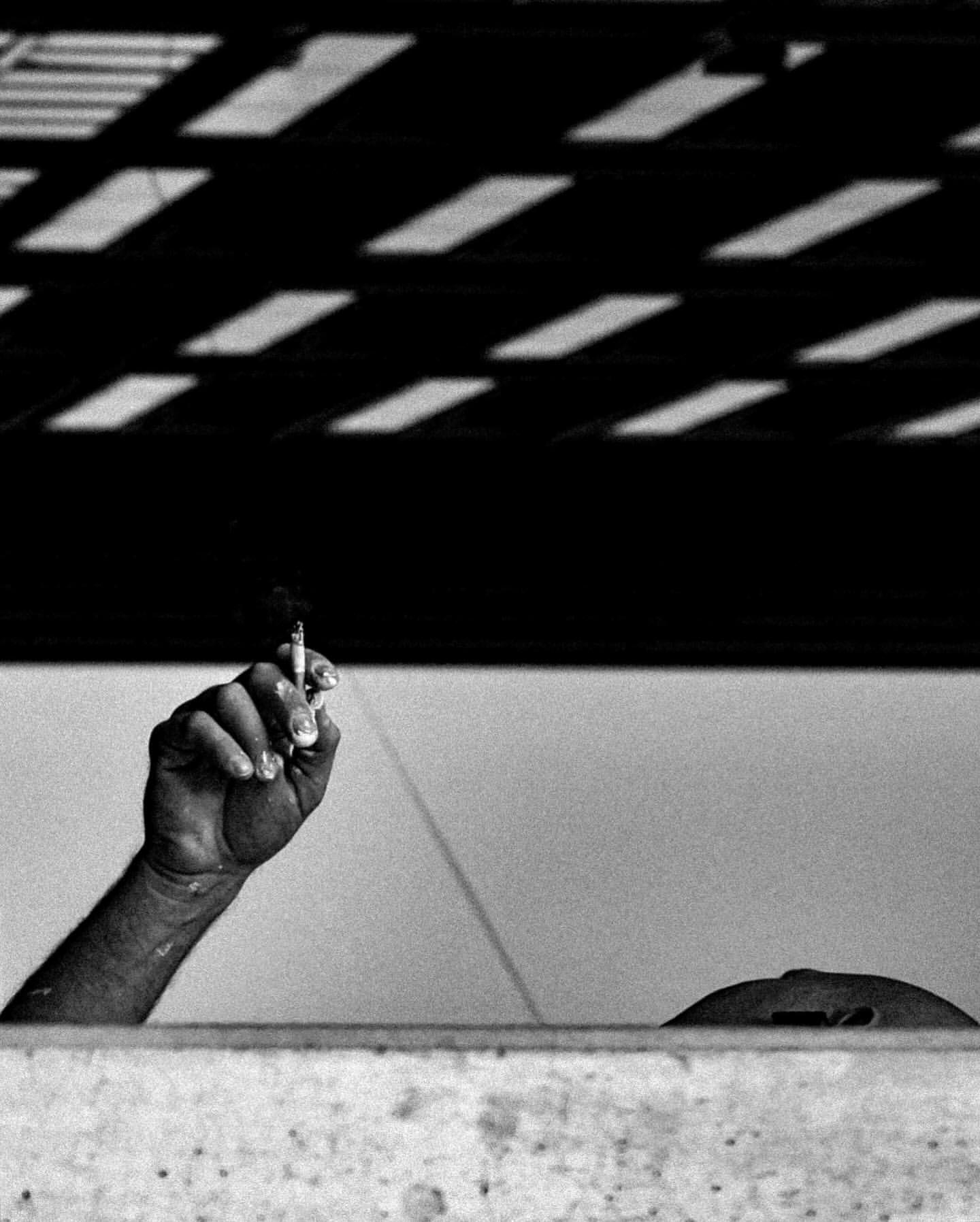Monochrome photograph of a person's hand holding a lit cigarette, with shadows creating abstract patterns in the background.