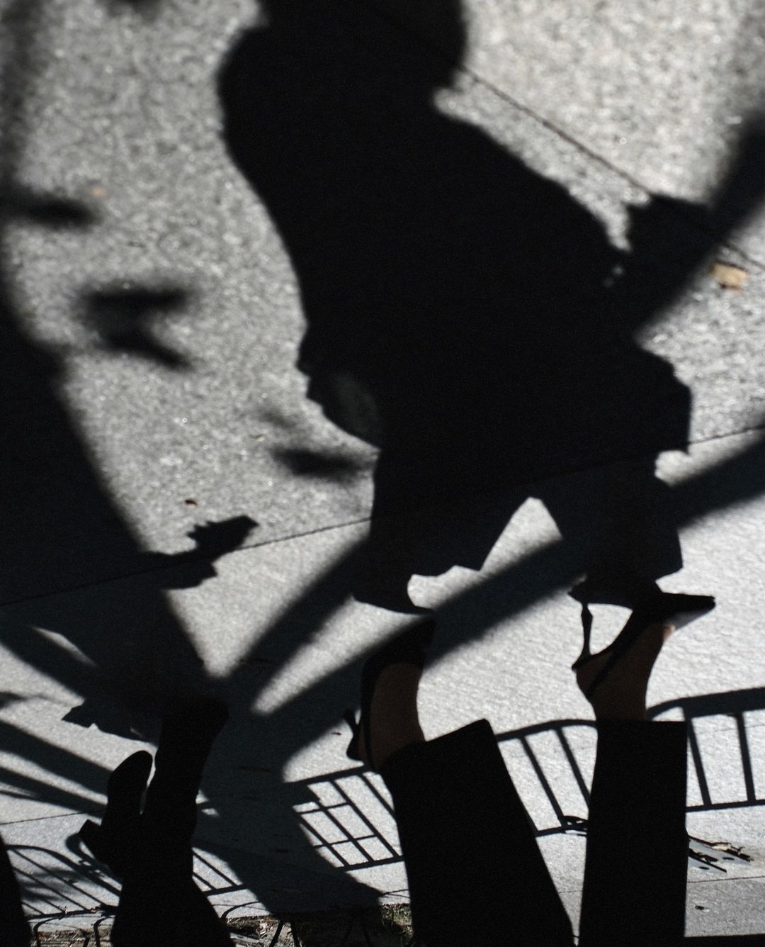 High-angle shot of pedestrian shadows and feet wearing heeled shoes on a concrete sidewalk, with hints of sunlight and trees.