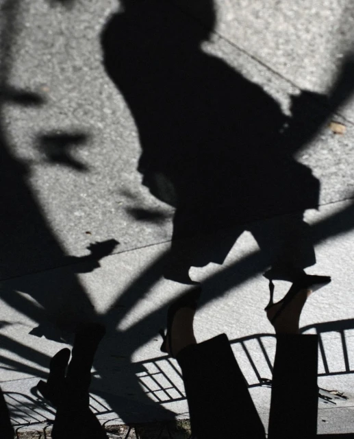 High-angle shot of pedestrian shadows and feet wearing heeled shoes on a concrete sidewalk, with hints of sunlight and trees.