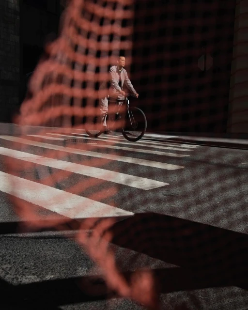 Man rides a bicycle across a crosswalk in an urban setting. Street-style fashion shoot with dappled light and shadow, dark tones.