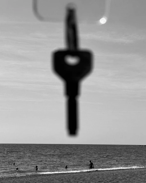 Black and white shot of a key hanging in the foreground with a beach and ocean scene with people in the background.