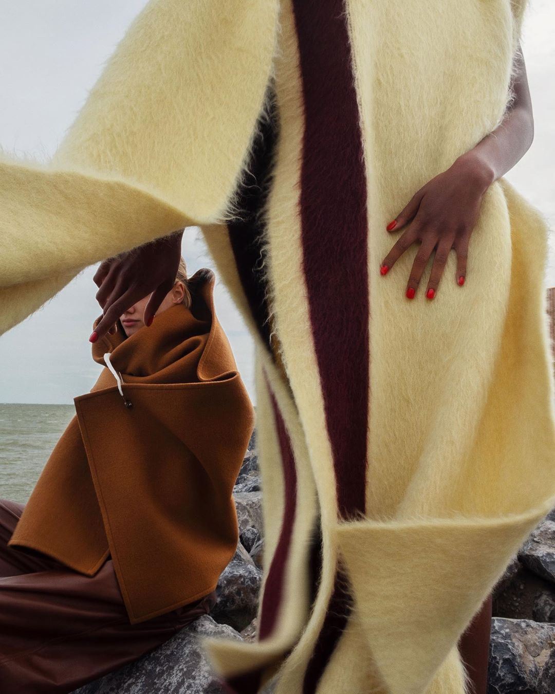 Two figures on a rocky beach wearing stylish coats in a fashion editorial. One person wears a striped, cream colored, wool shawl.