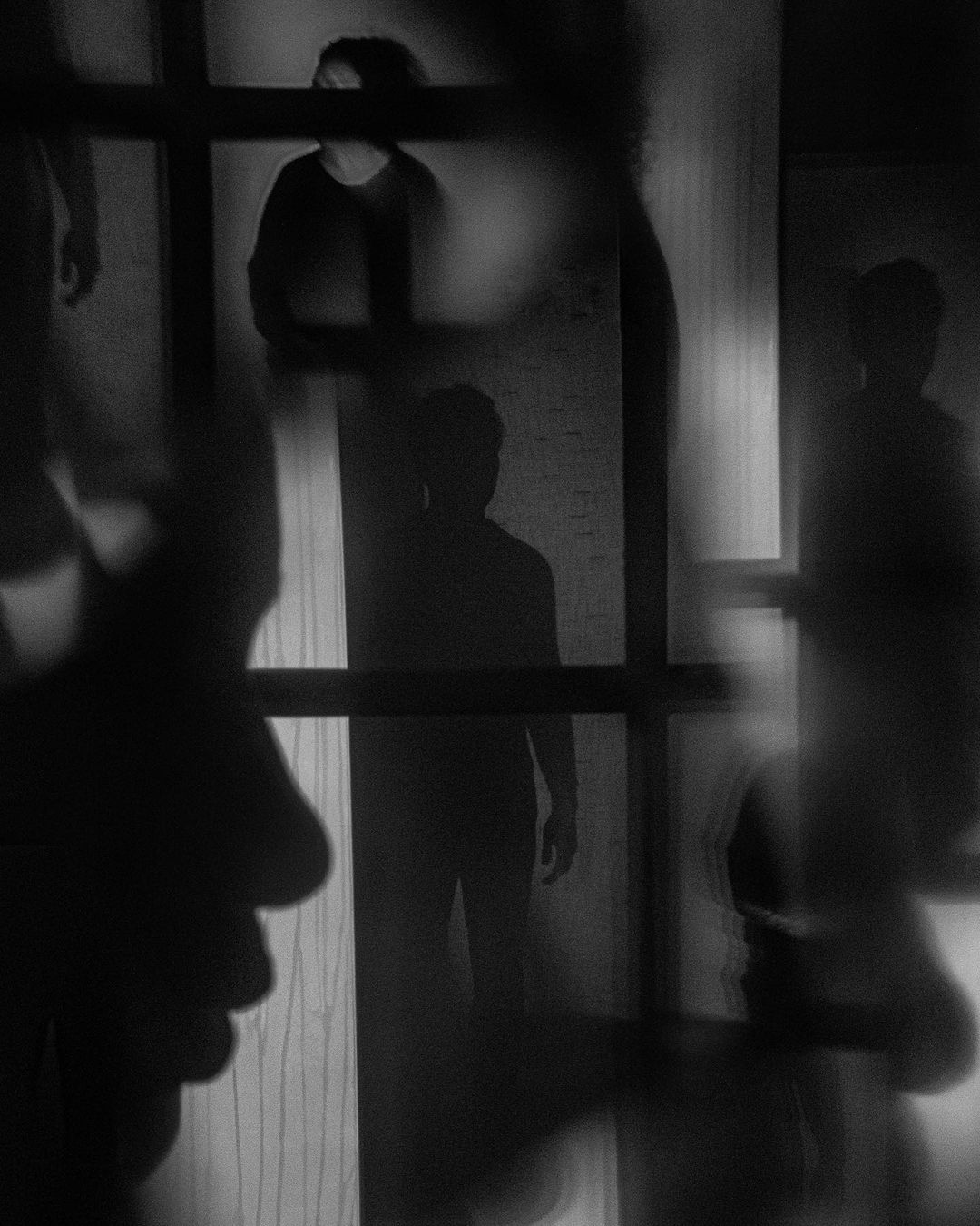 Monochromatic image featuring multiple figures silhouetted against a wall, viewed through a partially obscured window frame.