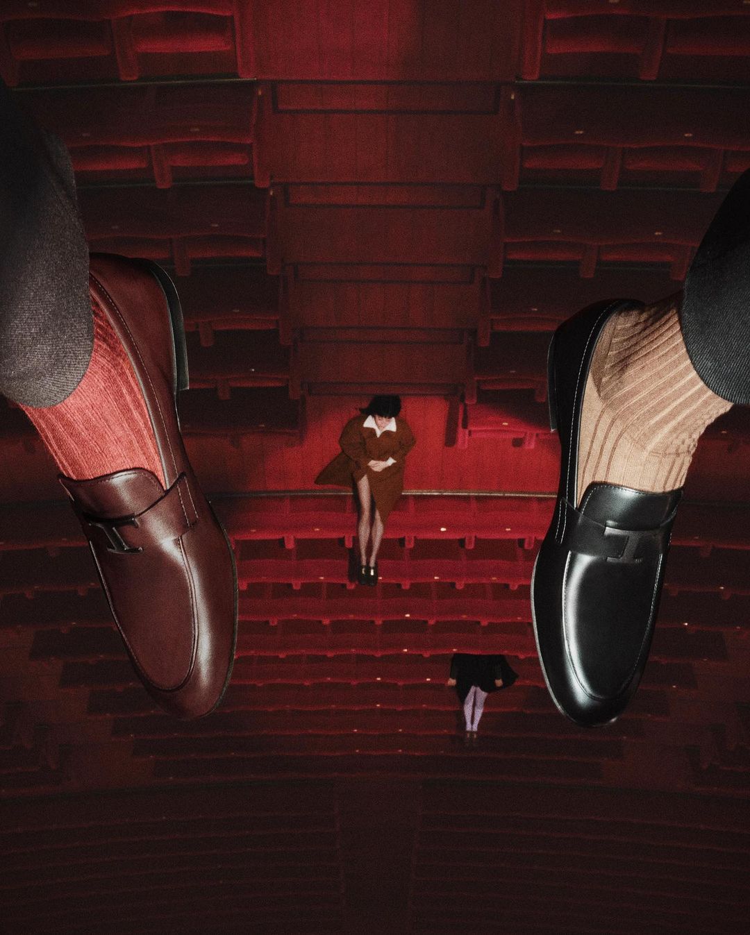 Eye-level, abstract composition of red theatre rows, two women standing on the steps, and two large men's dress shoes and socks.
