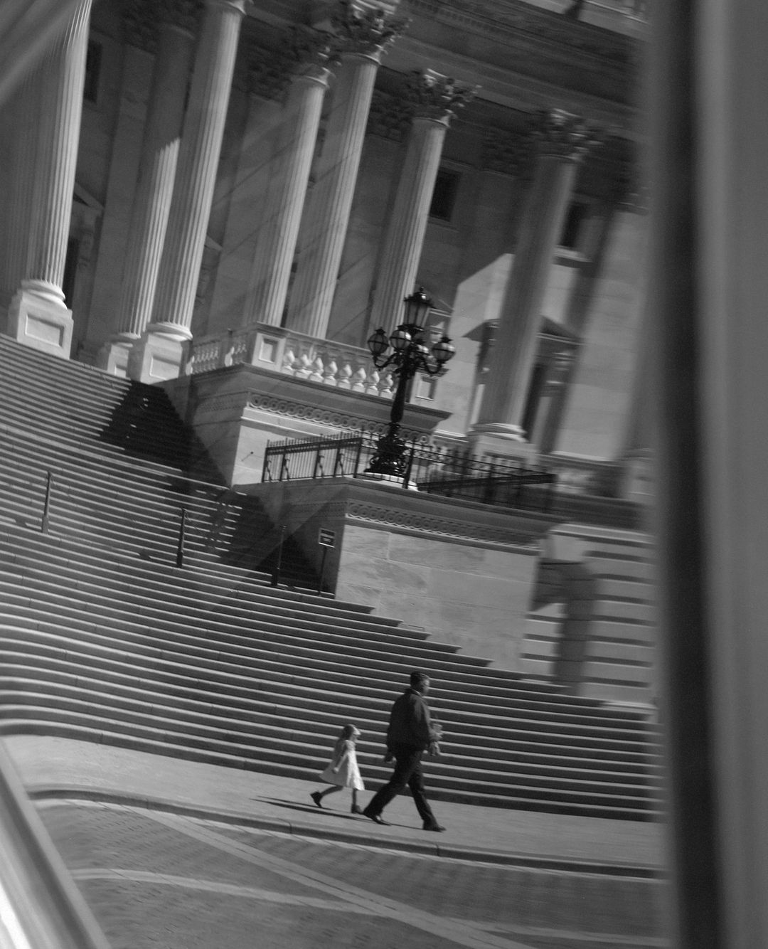 Black and white photo of an adult and a child walking on the steps of a building with large columns on a sunny day.