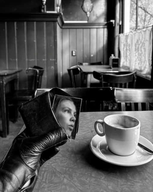 Monochromatic photo of a leather-gloved hand holding a wallet with a woman's image, juxtaposed with a cup of coffee in a cafe.