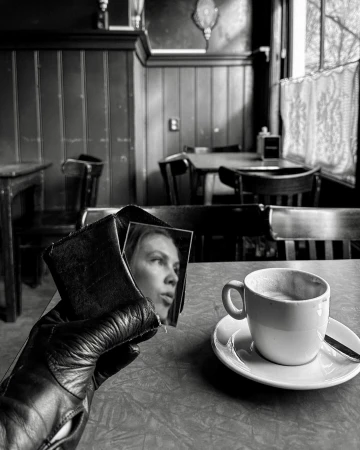 Monochromatic photo of a leather-gloved hand holding a wallet with a woman's image, juxtaposed with a cup of coffee in a cafe.