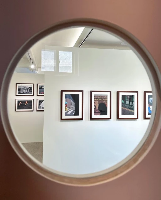 A view through a circular frame reveals an art gallery featuring framed photographs displayed on a white wall, illuminated by ceiling lights.