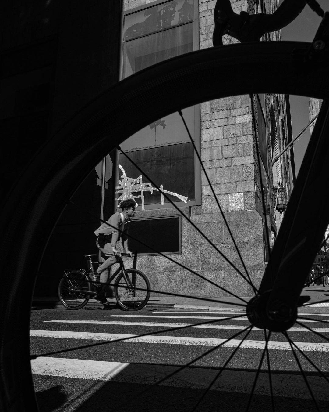 Monochrome shot of adult male riding a bicycle on a city street with crosswalk, viewed through a bicycle wheel in the foreground.