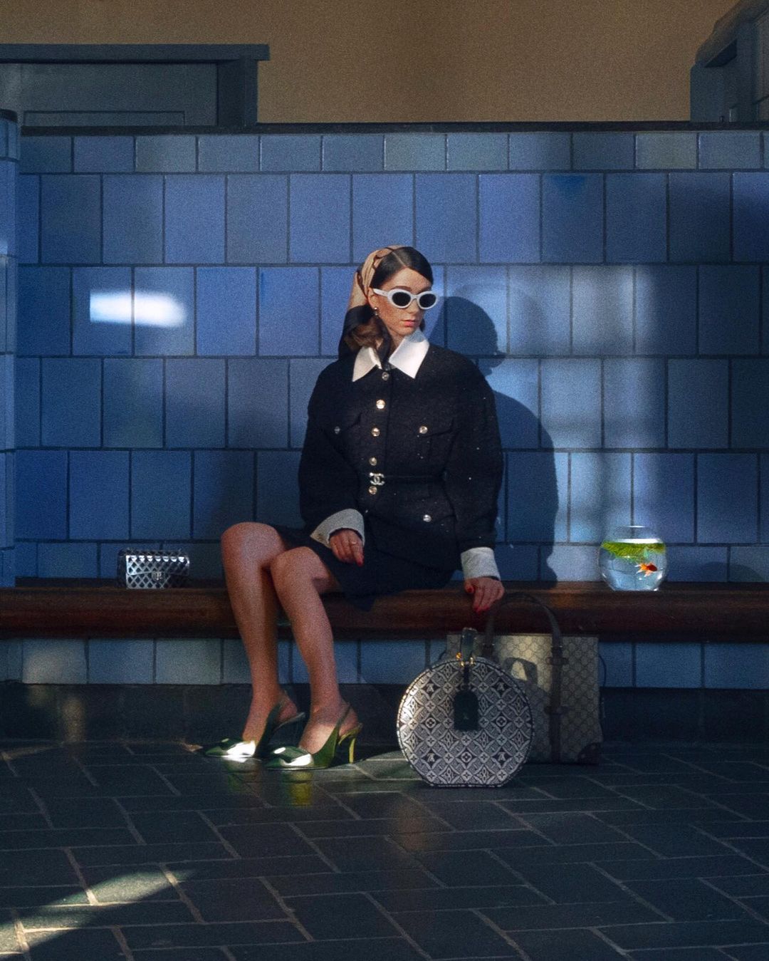 Woman in black dress and sunglasses sits on bench near blue tiled wall with purse, goldfish bowl, and coffee cup, casting shadow.