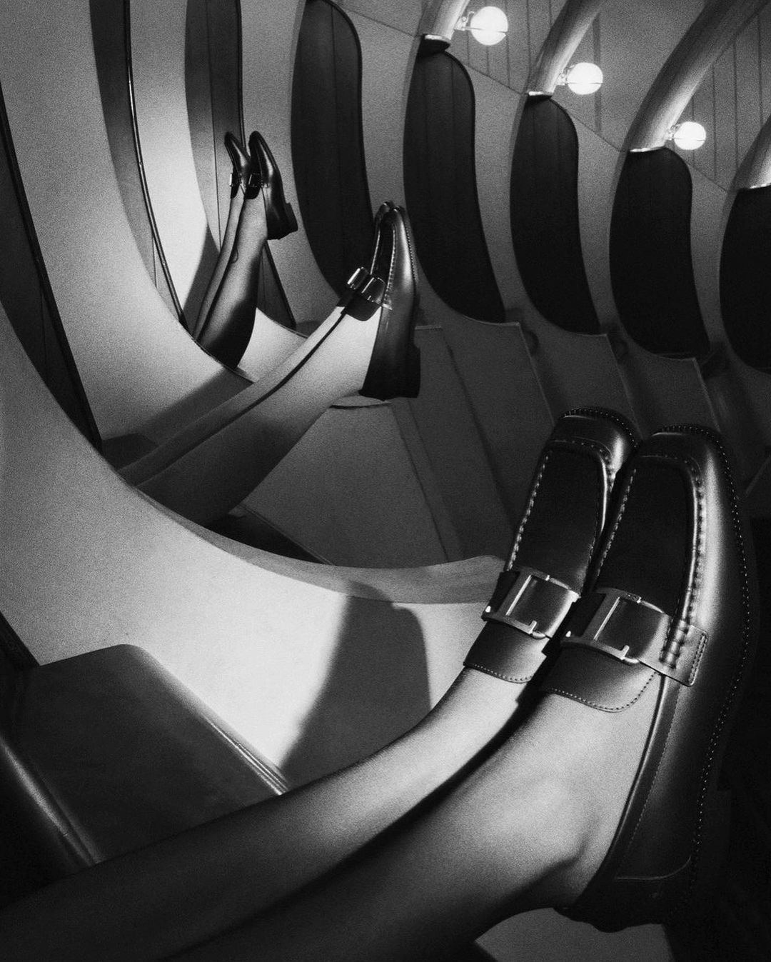Black and white fashion shot: A pair of loafers on human feet against a striped, curved background with bright, round lights.