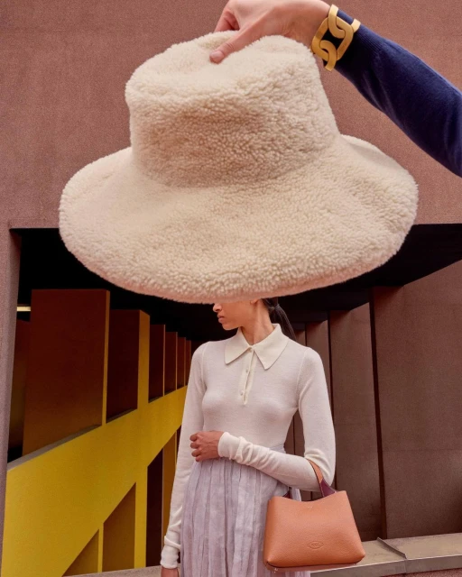 Fashion shot of a woman in a long-sleeve shirt and pleated skirt, holding a bag, standing beneath a large, fuzzy hat held by a hand.