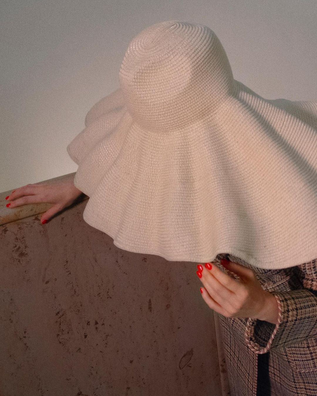 A woman wearing a large white sun hat and a plaid coat with red painted fingernails rests her hands on a marble ledge indoors.
