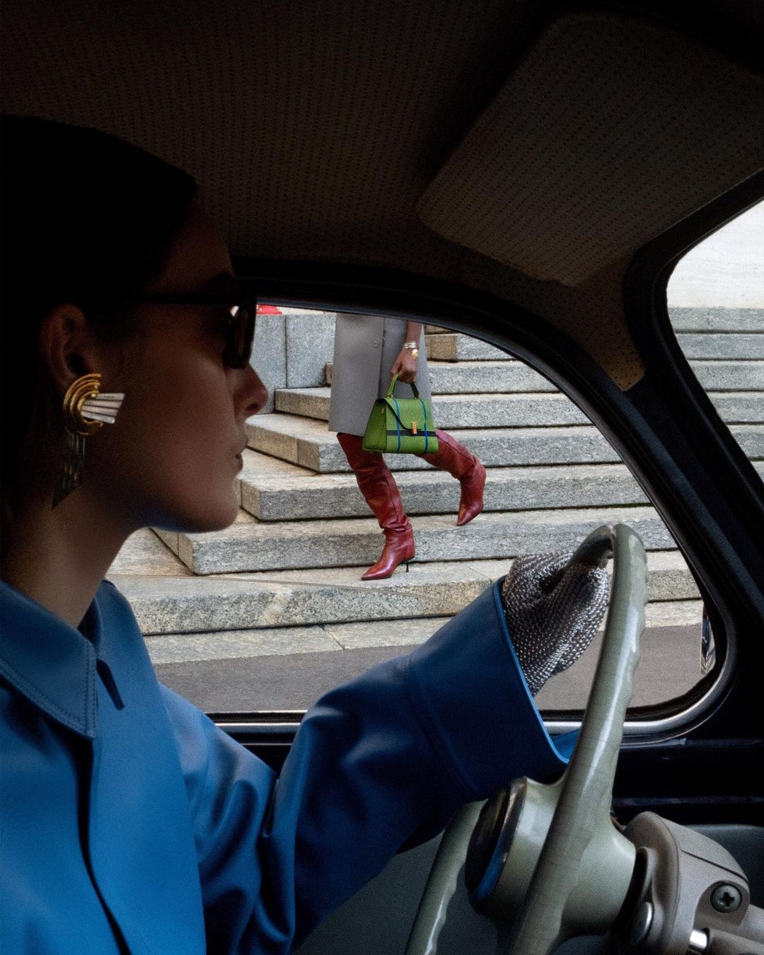 Woman driving vintage car while second woman in leather boots walks up steps with handbag in stylish outdoor fashion shot.