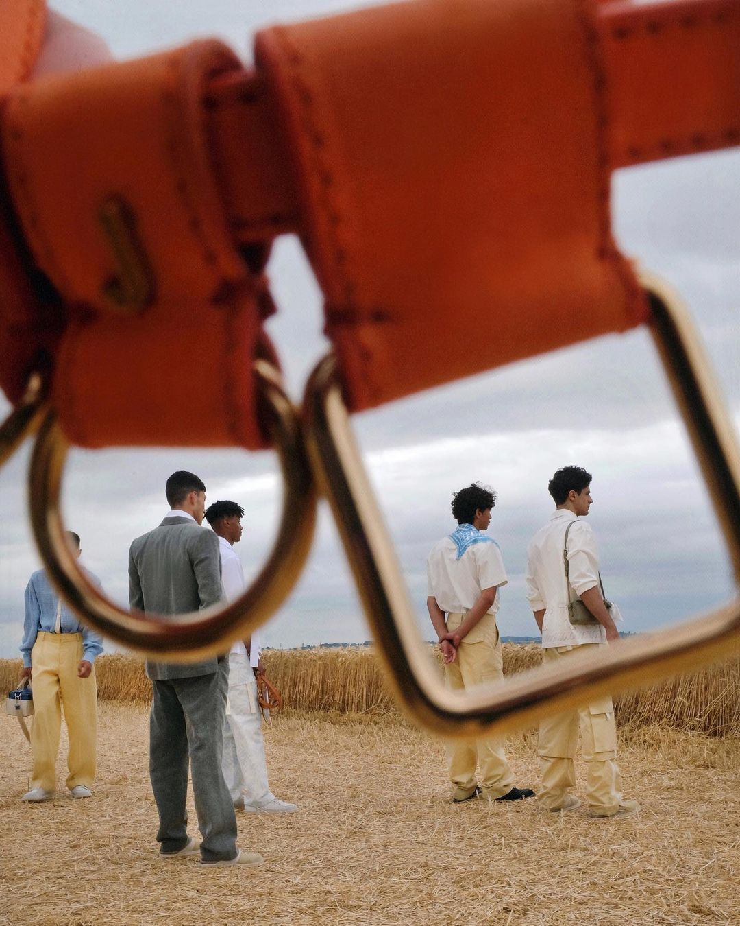 Group of men stand in a field, partially framed by an orange leather handbag strap with metal hardware. Cloudy sky background.