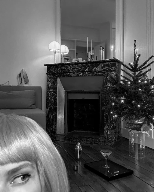 Black and white shot of a woman with blonde wig in an indoor setting with a fireplace, Christmas tree, and cocktail accessories.