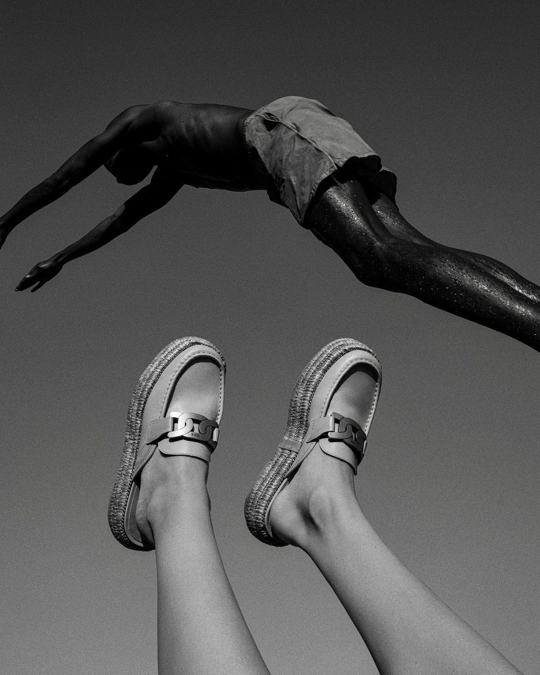 Monochromatic medium shot of a person diving above legs and shoes pointed upward against a stark grey background in bright sunlight.