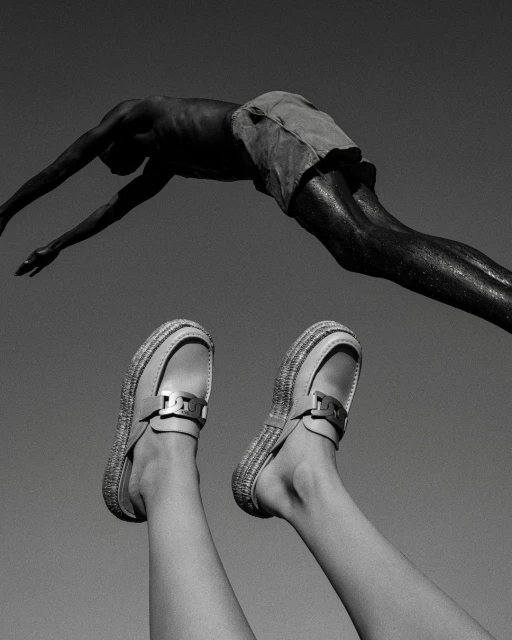 Monochromatic medium shot of a person diving above legs and shoes pointed upward against a stark grey background in bright sunlight.