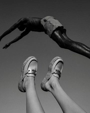 Monochromatic medium shot of a person diving above legs and shoes pointed upward against a stark grey background in bright sunlight.