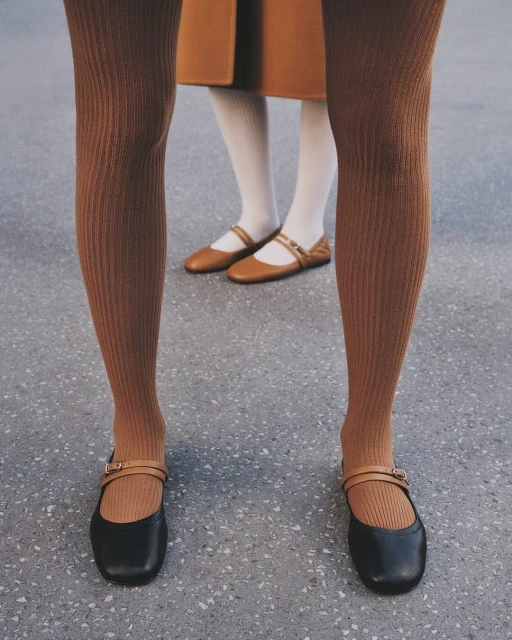 Close-up fashion shot of two women wearing tights and black and tan ballet flats standing on a grey concrete surface.