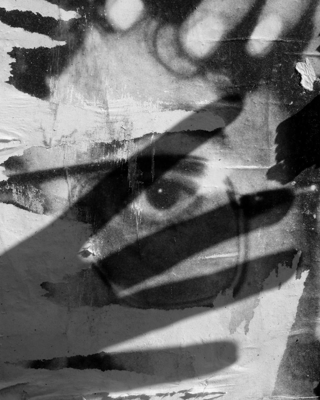 Monochrome image of a partial face and eye peeking out from behind strong linear shadows, resembling bars, against a textured background.