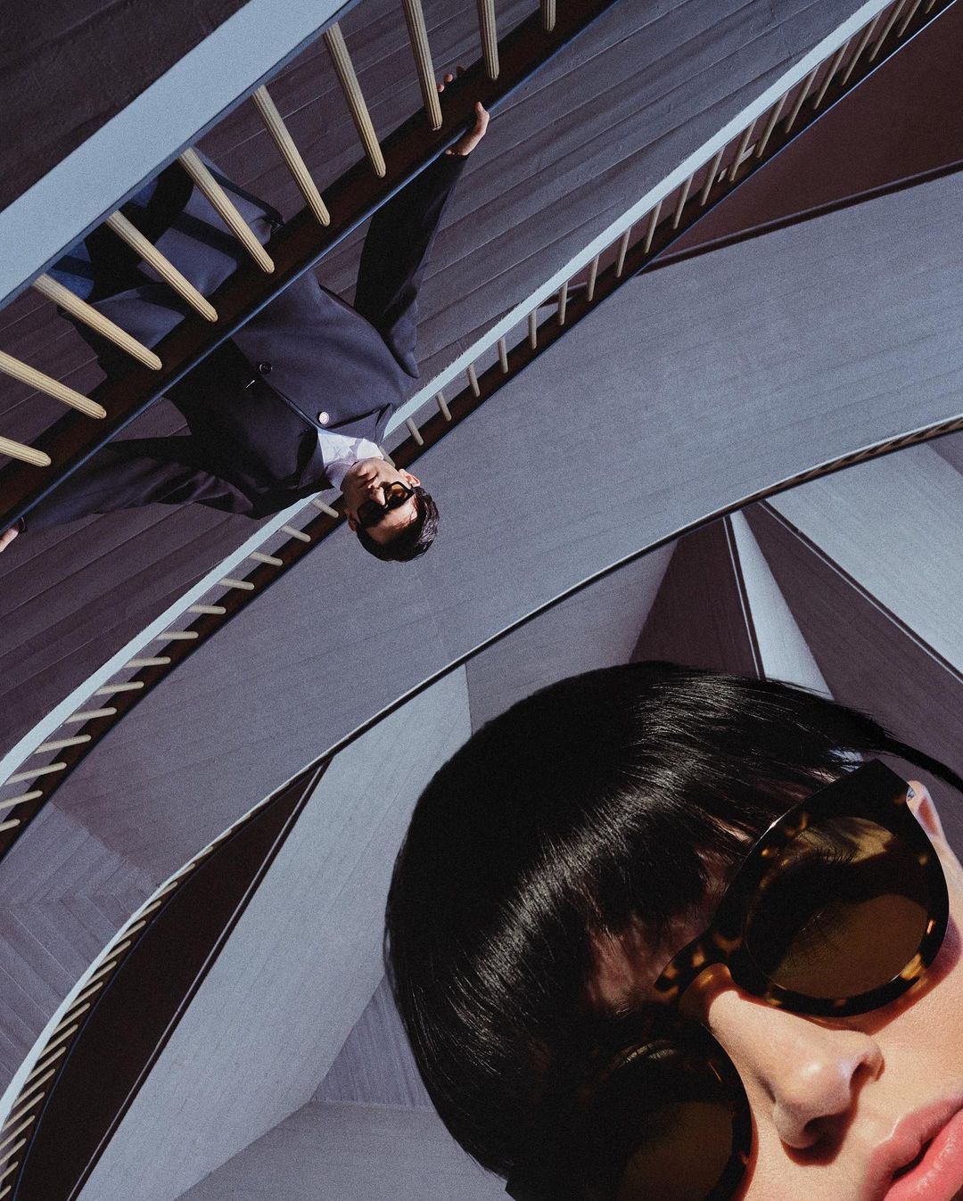 Fashion shot featuring a man in a suit on a ladder and a woman's face with sunglasses, set against a modern architectural backdrop.