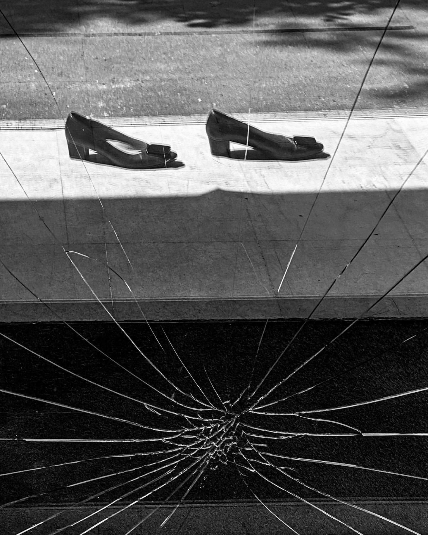 Two women's shoes are placed on a sidewalk, their reflections visible in the fractured glass of a nearby storefront window.