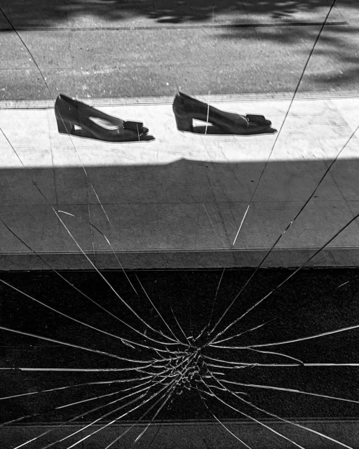 Two women's shoes are placed on a sidewalk, their reflections visible in the fractured glass of a nearby storefront window.