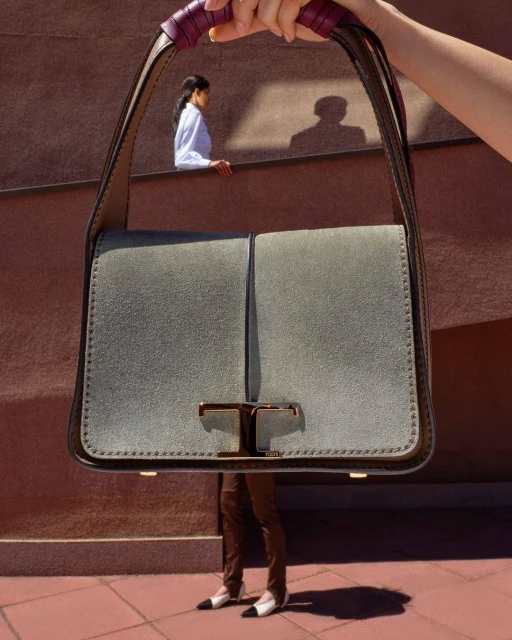 Leather handbag with double T logo in grey being held in front of an adult woman standing against a brown wall in an outdoor setting.
