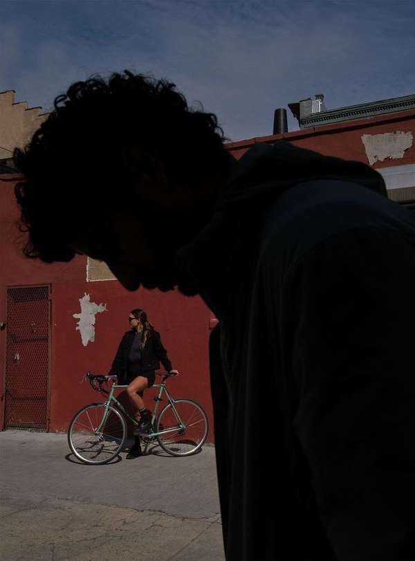 Woman sits on a bicycle in an alley, posing beside a red brick wall under a blue sky, with a blurred figure in the foreground.