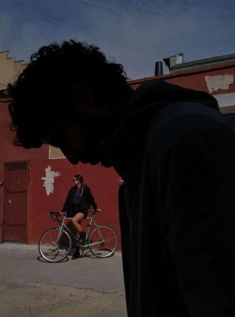 Woman sits on a bicycle in an alley, posing beside a red brick wall under a blue sky, with a blurred figure in the foreground.
