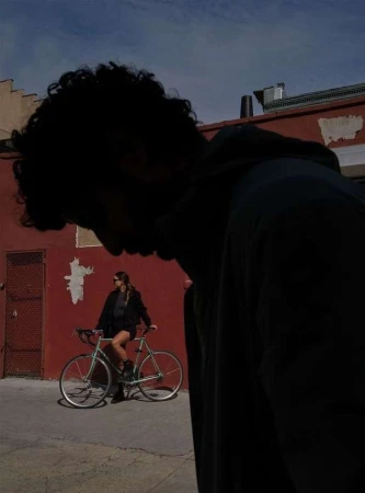 Woman sits on a bicycle in an alley, posing beside a red brick wall under a blue sky, with a blurred figure in the foreground.