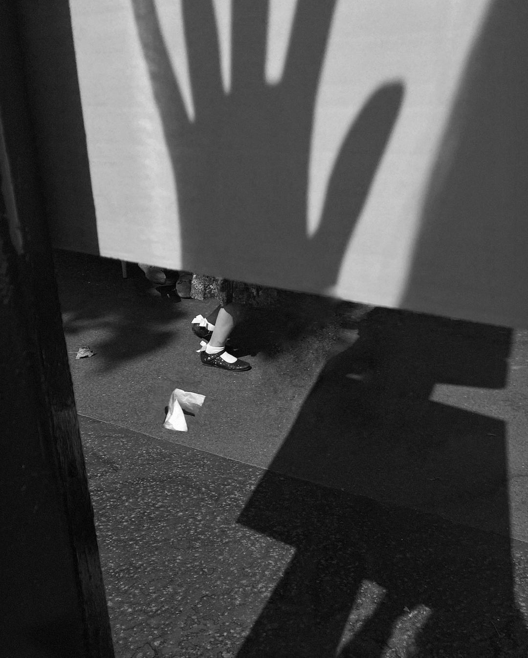 Black and white photograph of a child's feet in Mary Jane shoes, next to a crumpled white paper, and cast shadows of a hand and object.