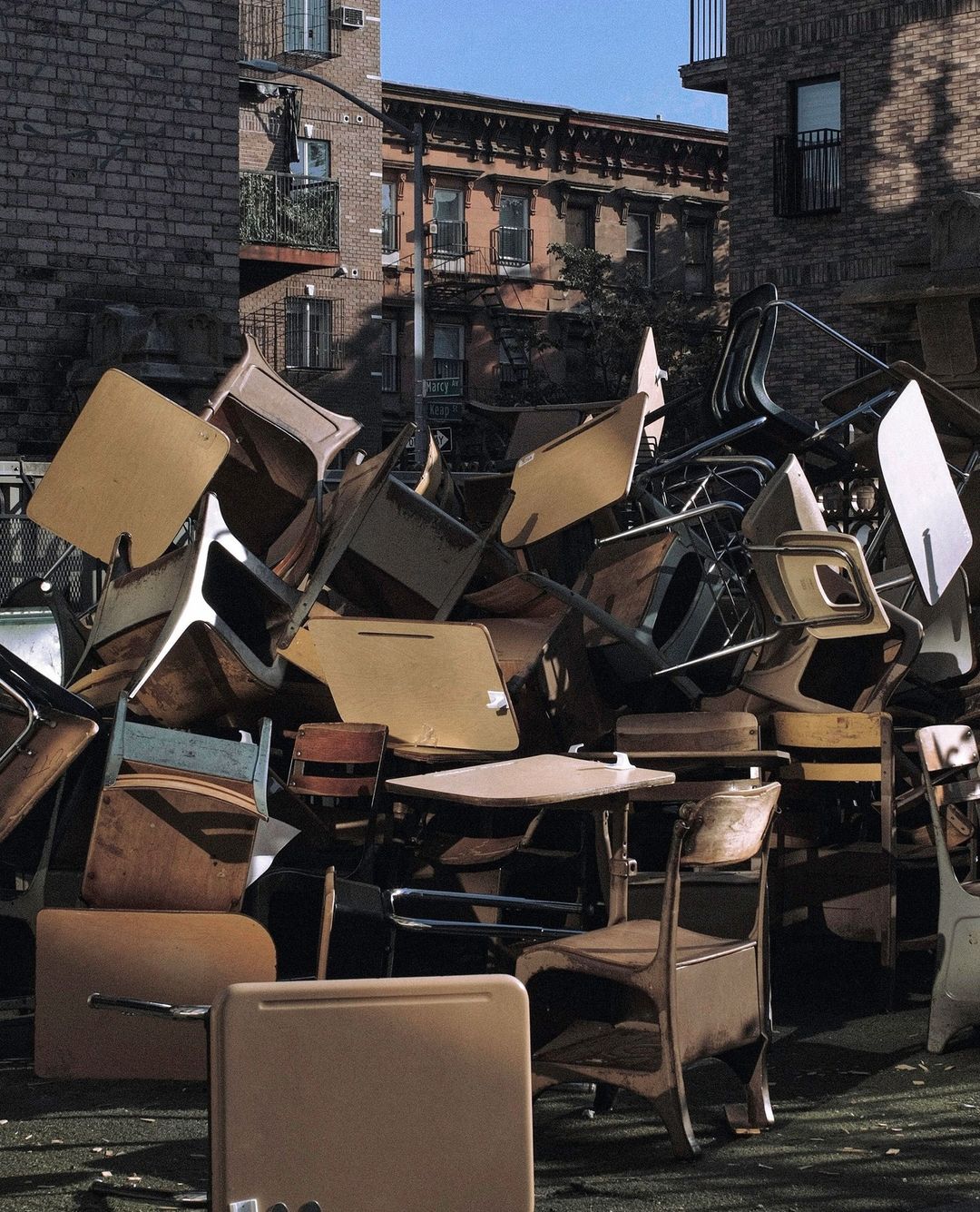 Pile of old school desks and chairs in an outdoor urban setting with brick buildings in the background, creating a scene of disposal.