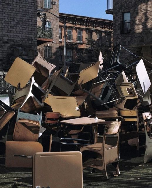 Pile of old school desks and chairs in an outdoor urban setting with brick buildings in the background, creating a scene of disposal.