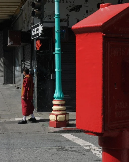 Urban scene: A monk walks along a city street past a vintage-style light post and red fire alarm box on a sunny day.