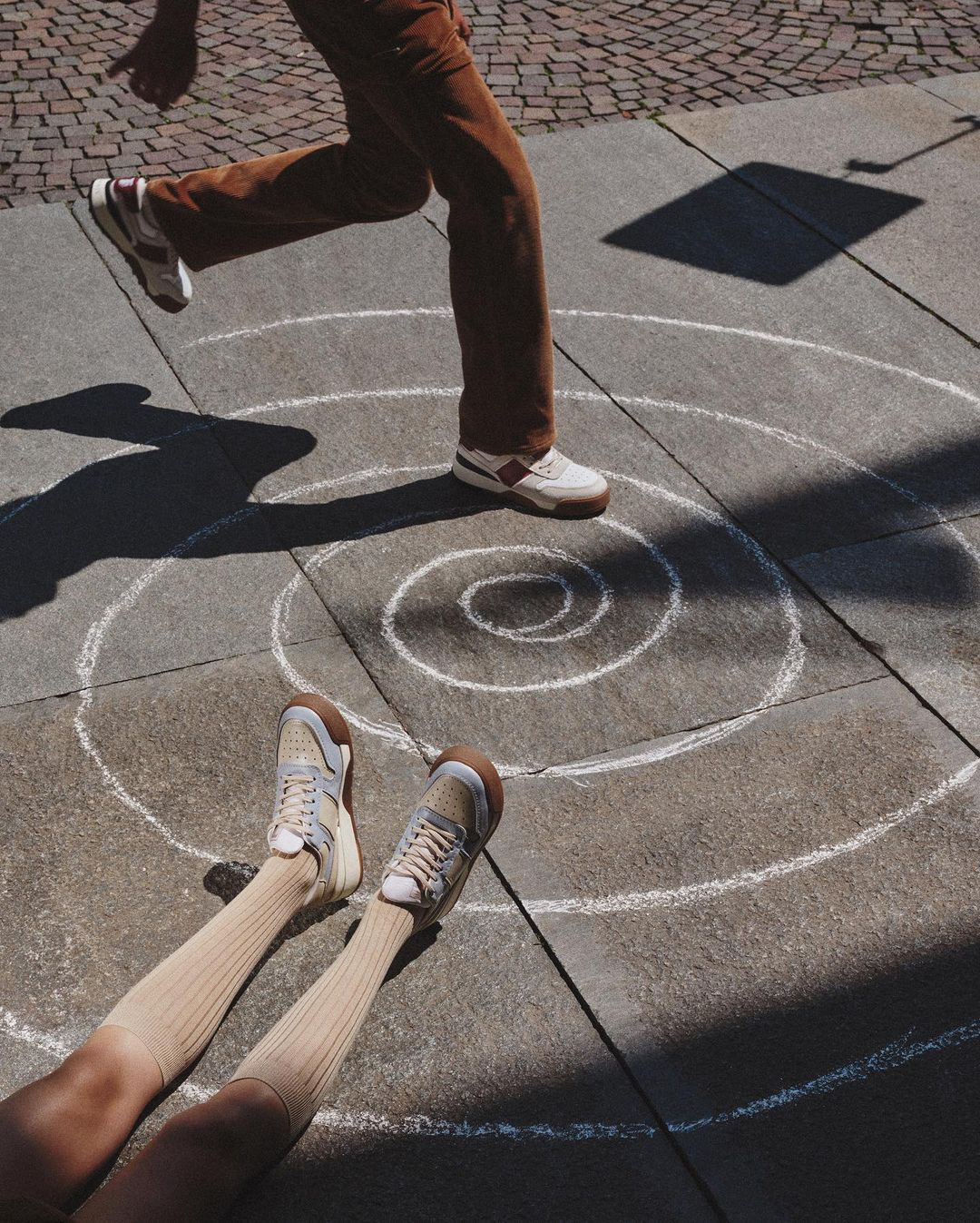 Person running in the sun with white sneakers in the street on a chalk-drawn concentric circle, person lying in the circle with sneakers.