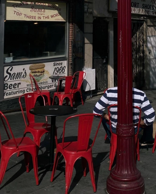Outdoor seating area in front of Sam's Burgers, featuring red metal chairs and black tables on a sidewalk, with a partially obscured person.
