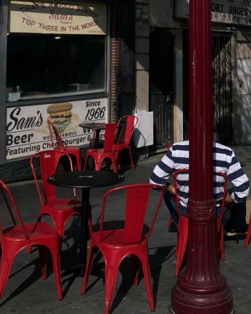 Outdoor seating area in front of Sam's Burgers, featuring red metal chairs and black tables on a sidewalk, with a partially obscured person.