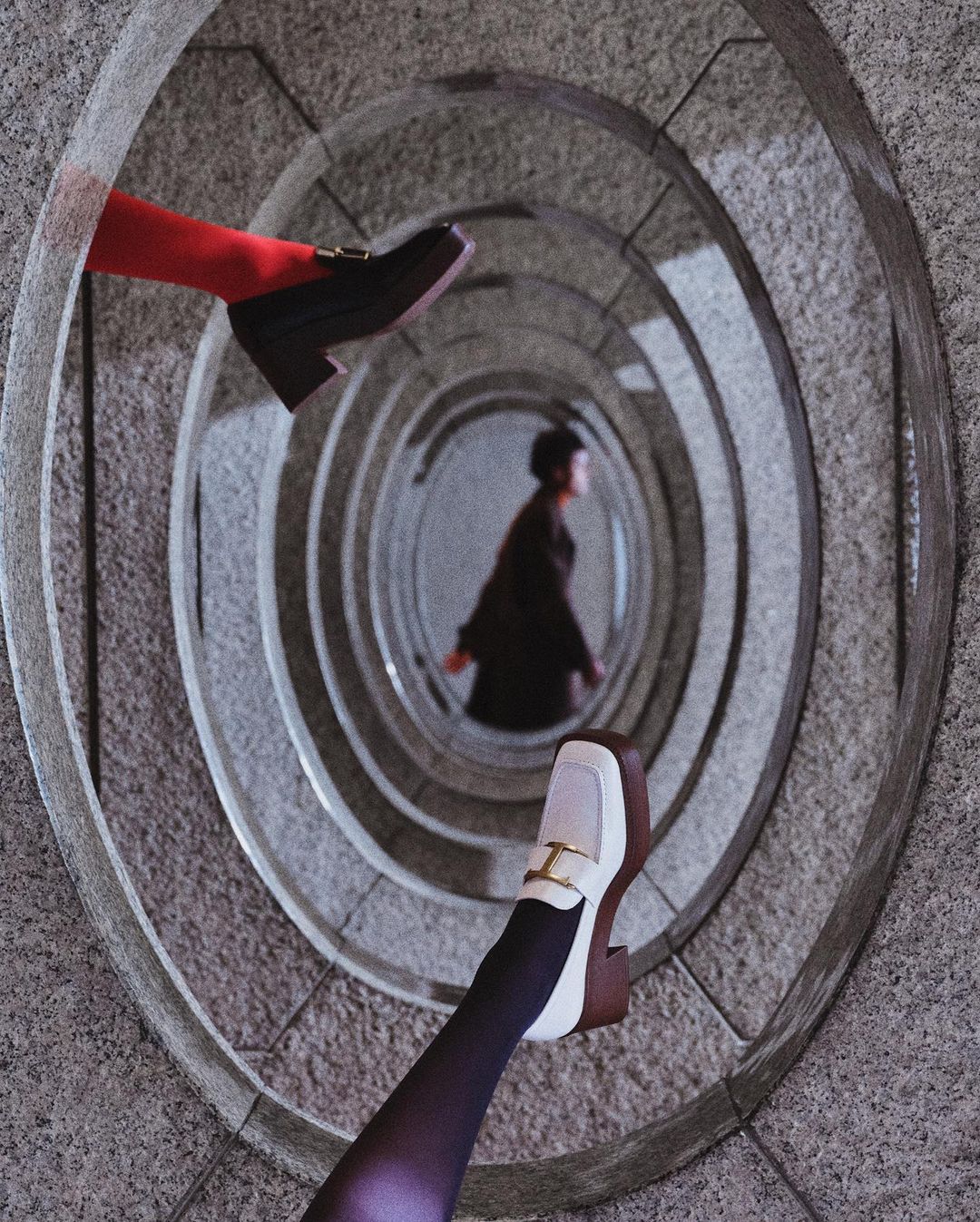 Eye-level angle of a stone spiral staircase, with two women; one is centered within the spiral, and the other's legs and shoes are visible.