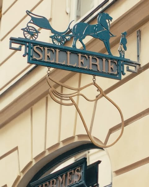 Metal sign for Sellerie featuring a horse-drawn carriage and a horse's head, mounted on a light-colored building.