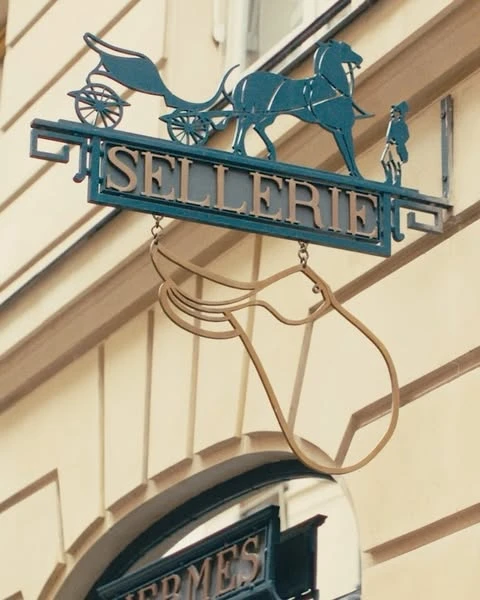 Metal sign for Sellerie featuring a horse-drawn carriage and a horse's head, mounted on a light-colored building.