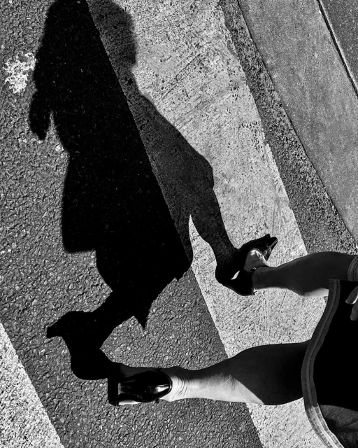 Black and white photo of a person's legs and feet in dress shoes walking on a crosswalk with their shadow cast in front of them.