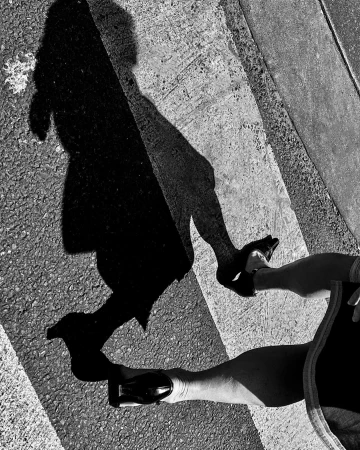 Black and white photo of a person's legs and feet in dress shoes walking on a crosswalk with their shadow cast in front of them.