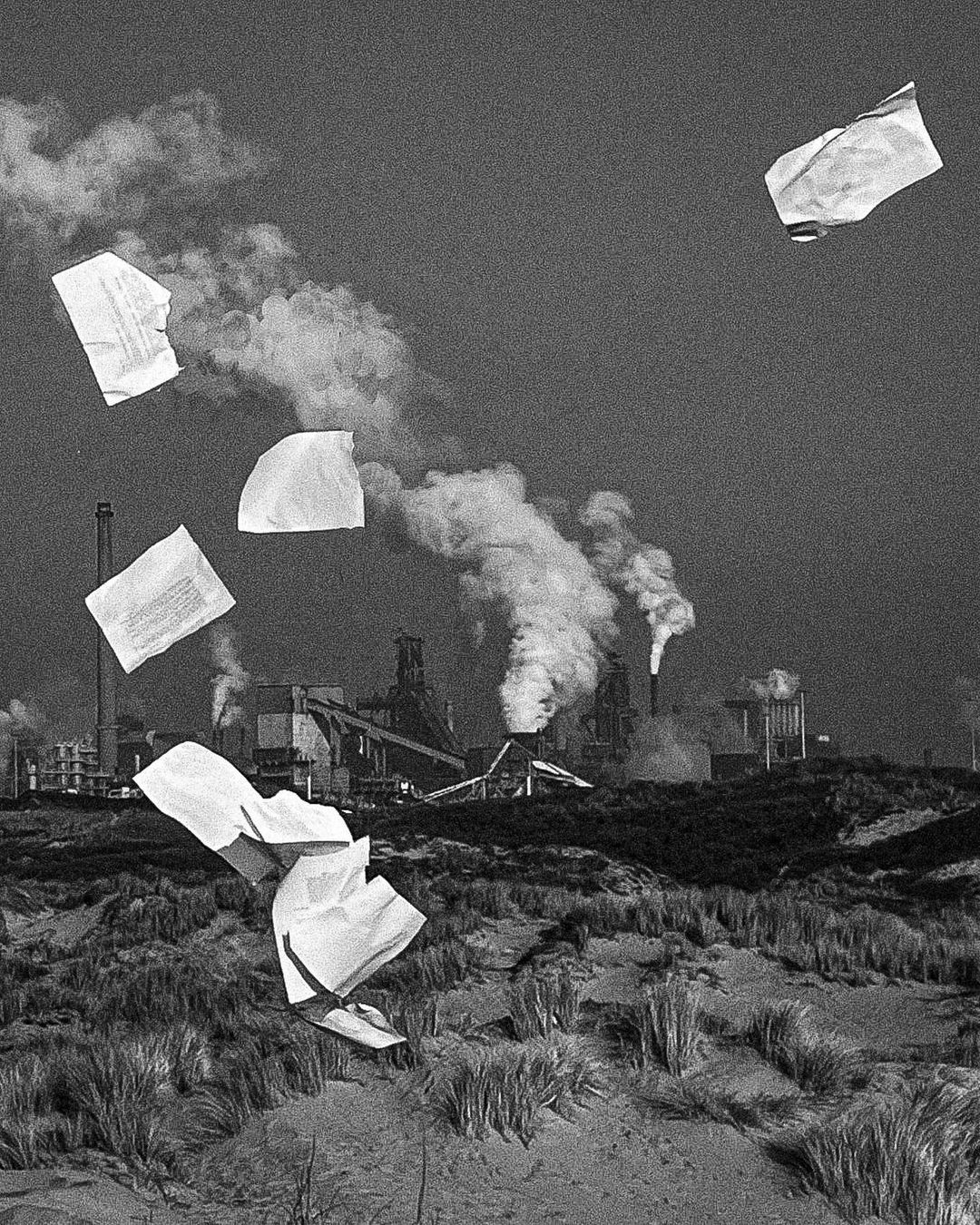 Black and white photo of papers blowing in the wind near dunes with an industrial plant and smokestacks in the background.