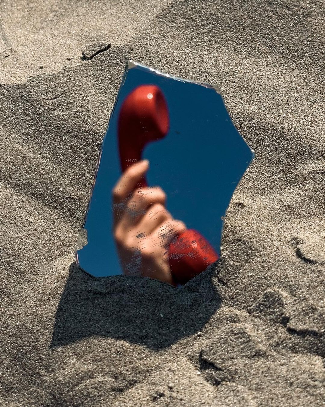 Reflection in a broken mirror shows a hand holding a red telephone receiver, partially buried in fine, grey sand, outdoors on a sunny day.