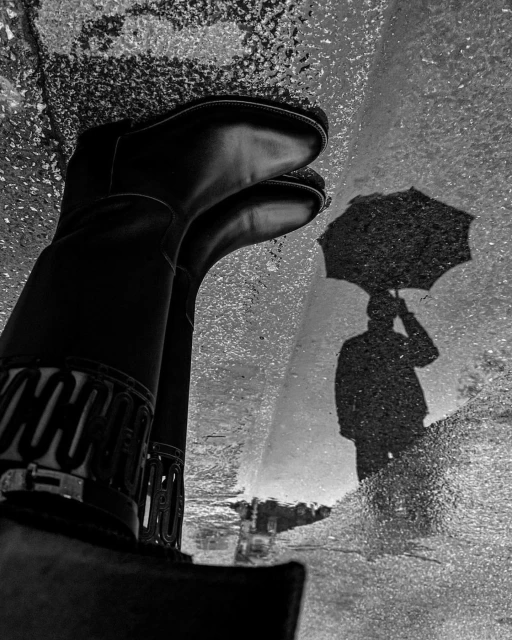 Black and white shot of black boots reflected in a puddle with the shadow of a person holding an umbrella on a wet city street.
