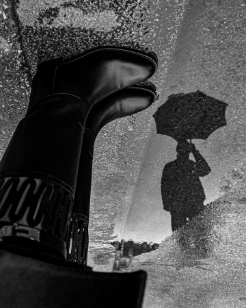 Black and white shot of black boots reflected in a puddle with the shadow of a person holding an umbrella on a wet city street.