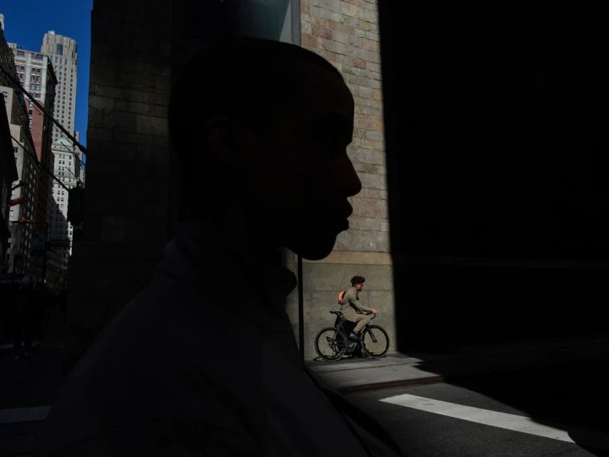 Silhouette of a person juxtaposed with a cyclist on a city street, highlighting contrasts of light and shadow near a building.