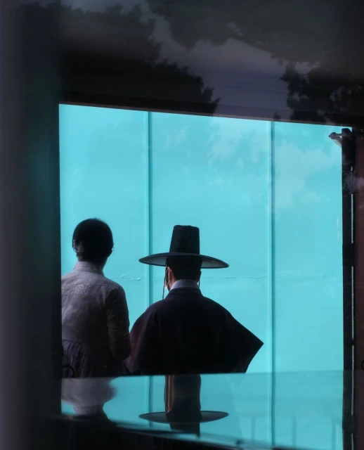 Two figures, one wearing a traditional Korean hat, stand at a window looking out onto a pale blue scene, partially framed by dark architectural elements.